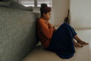 woman in orange long sleeve shirt sitting on the floor with her hands on her chest looking anxious